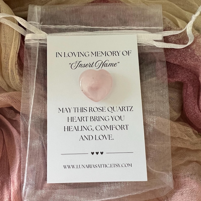 Op de afbeelding: Een kleine, transparante organzazak met een roze rozenkwartshart en een witte kaart. De kaart leest "IN LOVING MEMORY OF 'Insert Name'" en "MAY THIS ROSE QUARTZ HEART BRING YOU HEALING, COMFORT AND LOVE." Het websiteadres staat ook op de kaart.