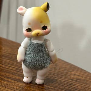 Puede incluir: Una pequeña muñeca hecha a mano con cara de oso y un pelele de punto gris. La muñeca tiene una cara blanca con detalles amarillos en la cabeza y las orejas, y mejillas rosadas. Está sobre una superficie de madera.