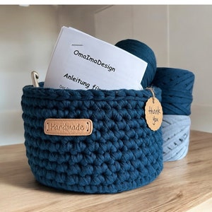 Könnte beinhalten: Ein handgefertigter, dunkelblauer Häkelkorb mit einem Lederetikett "Handmade". Der Korb enthält einen Häkelhaken, einen Knäuel tealfarbenes Garn und ein Heft. Zwei weitere Knäuel Garn in Blautönen befinden sich hinter dem Korb. Ein kleines Etikett sagt "Thank You".