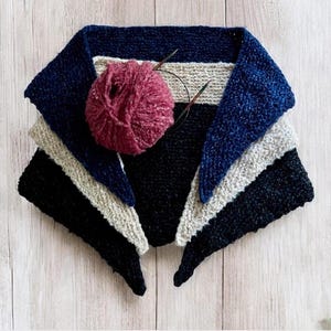 May include: A triangular knitted scarf in shades of navy blue, black, and beige. The scarf is folded in thirds and a ball of pink yarn is resting on top.