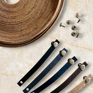 Könnte beinhalten: Eine Auswahl an Lederriemen in Schwarz, Marineblau, Braun und Kork, jeweils mit einem silbernen Knopfniet. Eine Rolle Korkmaterial und mehrere silberne Nieten sind ebenfalls sichtbar. Die Artikel sind auf einer Marmoroberfläche angeordnet.