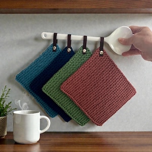 Könnte beinhalten: Vier quadratische, gestrickte Topflappen in Türkis, Marineblau, Grün und Mauve hängen an einer weißen Stange mit braunen Lederriemen. Eine weiße Tasse steht auf einer Holzoberfläche. Eine Hand hält die Stange.