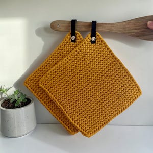 Könnte beinhalten: Zwei quadratische, senfgelbe Topflappen aus Strick mit schwarzen Lederaufhängeschlaufen. Die Topflappen hängen an einem Holzlöffel. Im Hintergrund befindet sich eine kleine Topfpflanze.