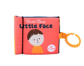 May include: A pink and blue soft book with a cartoon illustration of a boy's face. The book has a black strap and a yellow teething ring. The title of the book is "Fox and Bunny Little Face" and it says "For 1+ year".