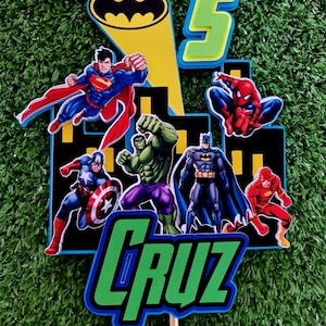 May include: A green and blue cake topper with a cityscape backdrop featuring various superheroes including Superman, Batman, Spiderman, Hulk, Captain America, and The Flash. The number 5 is on the top left corner and the name "CRUZ" is at the bottom.