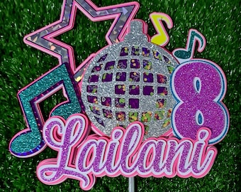 Può includere: Un topper per torta rosa e viola con una palla da discoteca argentata, note musicali, una stella e il nome "Lailani" con il numero 8.