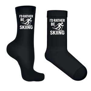 Może przedstawiać: Dwie czarne skarpetki z białym napisem "I'd Rather Be Skiing". Skarpetki mają grafikę narciarza. Skarpetki są wykonane z miękkiego materiału i są odpowiednie dla entuzjastów narciarstwa.