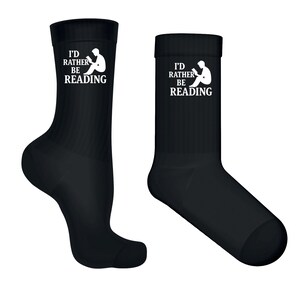 Op de afbeelding: Twee zwarte sokken met de tekst "I'D RATHER BE READING" in het wit. De tekst staat boven een witte afbeelding van een lezende persoon. De sokken zijn enkellang en gemaakt van zacht materiaal.