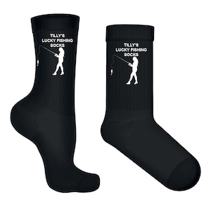 Puede incluir: Un par de calcetines negros de caña media con un estampado gráfico blanco de una persona pescando con caña y sedal. El texto "TILLY'S LUCKY FISHING SOCKS" está impreso en letras mayúsculas blancas sobre el gráfico en la parte superior.
