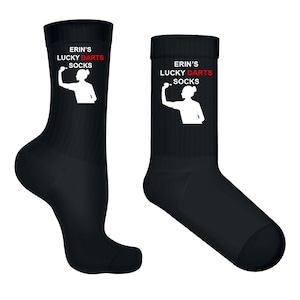 Puede incluir: Un par de calcetines negros tipo crew con un gráfico de silueta blanca de una persona lanzando un dardo. El texto "ERIN'S LUCKY DARTS SOCKS" está impreso en letras blancas y rojas sobre el gráfico en la parte superior de cada calcetín.