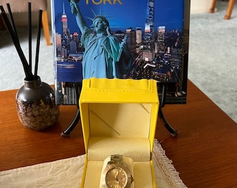 Puede incluir: Un reloj de pulsera Invicta de tono dorado en una caja amarilla. La caja tiene el logotipo de Invicta en la parte delantera y el reloj se muestra en el interior.
