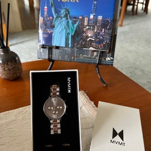 May include: A silver and rose gold watch with a chronograph face in a white box. The box has the brand name MVMT printed on it. The watch is sitting on a wooden table next to a book about New York City.