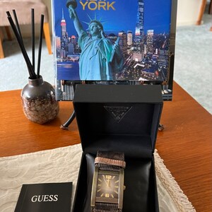 May include: A brown leather strap watch with a gold square face and a black box. The watch face has gold hands and gold numbers. The box has the word "GUESS" on it.