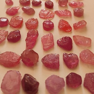 Puede incluir: Una colección de gemas en bruto, sin cortar, en tonos rosados y rojizos. Las piedras varían en tamaño y forma, con una textura rugosa y natural. Las gemas están dispuestas sobre una superficie clara, mostrando su belleza natural.