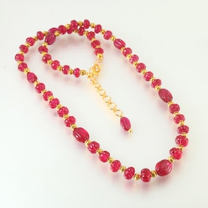 May include: A necklace featuring a strand of red, oval-shaped beads and smaller, round beads. The beads are interspersed with gold-colored spacers and connected by a gold chain. The necklace is displayed on a white surface.