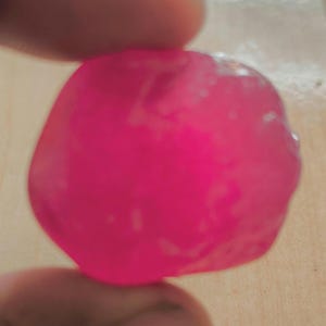 May include: A vibrant, rounded, pink object is held between two fingers. The object has a smooth, slightly translucent appearance. The lighting creates highlights and shadows on its surface, giving it a three-dimensional look.