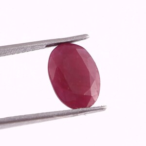 May include: A deep red oval-shaped gemstone with a faceted cut. The gemstone is held in place by a pair of tweezers.