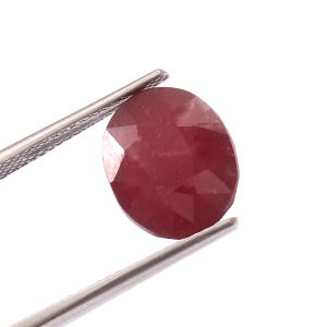 May include: A red oval gemstone with a faceted cut. The gemstone is held by a pair of tweezers.