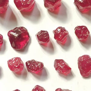 Könnte beinhalten: Eine Sammlung roher, ungeschliffener Rubin-Edelsteine. Die Steine haben eine tiefe, transparente rote Farbe, unregelmäßige Formen und eine raue Textur. Sie sind auf einer weißen Oberfläche angeordnet.