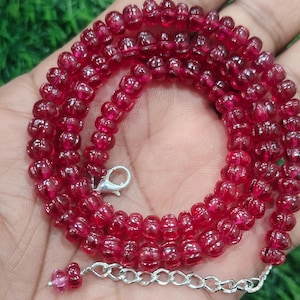 May include: A vibrant red beaded necklace with a silver clasp and extender chain. The necklace is composed of small, round, translucent beads. The beads are a deep, rich red color, and the necklace is displayed against a green background.