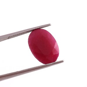 May include: A deep red oval gemstone held by tweezers. The gemstone is faceted and has a smooth, polished surface.