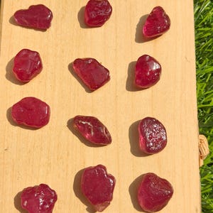 May include: A collection of raw, polished ruby gemstones displayed on a wooden board. The rubies vary in size and shape, with a deep red color and a glossy finish. The board is set against a backdrop of green grass.