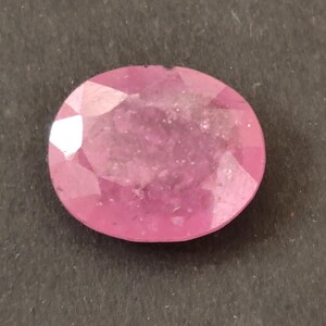Puede incluir: Una piedra preciosa rosa de forma ovalada y tallada, con una apariencia translúcida e inclusiones internas visibles. La piedra presenta una superficie pulida con múltiples facetas geométricas que reflejan la luz, mostrando diversos tonos de rosa vibrante.