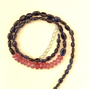 May include: A necklace featuring dark blue oval beads and faceted pink beads. The necklace has a silver chain and clasp. The beads are arranged in a spiral pattern against a cream background.