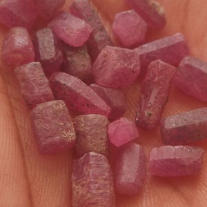 May include: A collection of rough, uncut ruby gemstones in varying shapes and sizes. The rubies display a range of pink and red hues, with some showing a slightly translucent quality. The gemstones are held in a hand, creating a natural presentation.