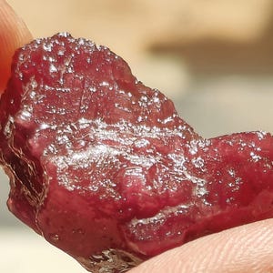 May include: A raw, deep red gemstone with a textured, crystalline surface. The stone features irregular facets and a glossy, reflective appearance, showcasing a rich crimson hue throughout its jagged, organic shape and uneven, multifaceted structure.