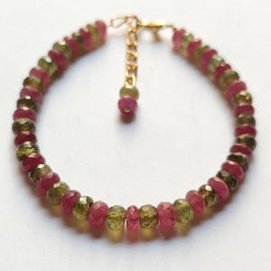 May include: A beaded bracelet featuring alternating ruby red and olive green gemstone beads. The bracelet is connected by a gold-colored chain and clasp. The gemstones are faceted, reflecting light. The bracelet is designed for wrist wear.