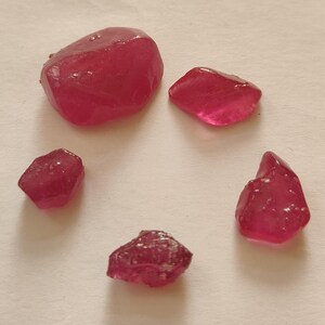 May include: Five loose, raw, deep pinkish-red gemstones of varying sizes and irregular, angular shapes. Each stone displays a translucent quality with a polished, faceted appearance, showcasing natural textures and vibrant, saturated colour tones.