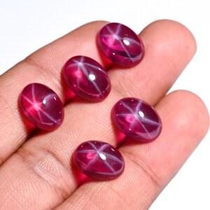 May include: Five oval-shaped, red star ruby cabochons with a starburst pattern. The cabochons are set on a hand.