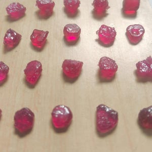 Puede incluir: Una colección de gemas de rubí en bruto, cada una con un vibrante tono rojo. Las gemas están dispuestas sobre una superficie de madera clara, mostrando sus formas irregulares y texturas naturales. Los rubíes varían en tamaño y claridad, reflejando la luz.