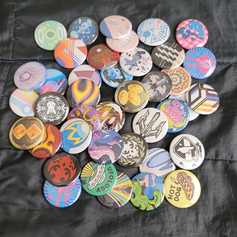May include: A collection of colorful round pins with various designs, including abstract patterns, animals, and text. The pins are arranged in a circle on a black surface.