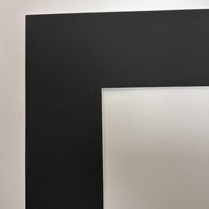 May include: A rectangular picture frame mount featuring a wide, solid black border surrounding a clean, white inner rectangular opening. The mount board has a smooth, matte finish with sharp, clean-cut edges defining the contrast between colours.