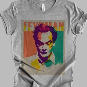 May include: A grey t-shirt with a vintage-style graphic of physicist Richard Feynman. The graphic features a colorful portrait of Feynman with the text "FEYNMAN" in a bold, retro font.