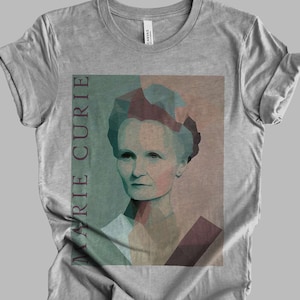May include: A gray t-shirt with a geometric portrait of Marie Curie in shades of green, blue, and pink. The text "Marie Curie" is printed vertically on the left side of the image.