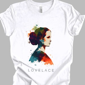 May include: White t-shirt featuring a colourful watercolour portrait of a woman with the text "LOVELACE" printed below.