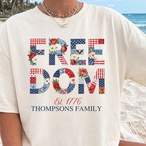 Puede incluir: Camiseta de color marfil con la palabra "FREEDOM" en un diseño de patchwork de patrones rojos, blancos y azules y acentos florales. El texto "Est. 1776 THOMPSONS FAMILY" está debajo de la palabra "DOM". La camiseta se lleva en la playa.
