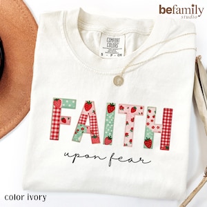 Puede incluir: Una camiseta de color marfil con la palabra FAITH en letras grandes de estilo bloque con un diseño de patchwork de cuadros vichy rojos, lunares verdes y patrones de fresas. Debajo de la palabra FAITH, la frase upon fear está en cursiva negra.