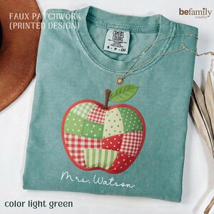 Puede incluir: Una camiseta verde claro con un diseño de manzana en falso patchwork con patrones rojos, verdes y blancos. El gráfico de la manzana incluye una hoja verde y un tallo marrón. Debajo de la manzana, el texto dice Mrs. Watson en letra cursiva.