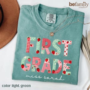 Puede incluir: Una camiseta verde claro con el texto FIRST GRADE en estilo patchwork con patrones de cuadros rojos, flores rosas y lunares verdes. Pequeñas ilustraciones de fresas están impresas en las letras. El nombre miss sarah aparece debajo.