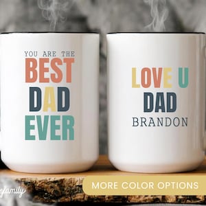 May include: Two white ceramic mugs with black handles and rims. One mug reads "YOU ARE THE BEST DAD EVER" in colorful text. The other mug says "LOVE U DAD BRANDON" in similar colors. The mugs are on a wooden surface.