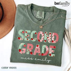 Puede incluir: Una camiseta de manga corta color verde musgo con el texto SECOND GRADE en una mezcla de patrones de cuadros vichy rojos y lunares verde claro con gráficos de fresas. El nombre miss emily está impreso en letra blanca.