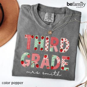 Puede incluir: Una camiseta color pimienta con el texto THIRD GRADE en letras con patrones de cuadros rojos, lunares verdes y fresas. Debajo del texto, el nombre mrs. smith está impreso en letra cursiva blanca. La camiseta tiene un cuello redondo.