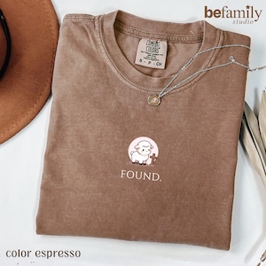 Puede incluir: Una camiseta de cuello redondo marrón con un pequeño gráfico de un cordero blanco de pie junto a una cruz marrón dentro de un círculo rosa claro, con la palabra FOUND. impresa en blanco debajo del gráfico. La camiseta tiene una etiqueta Comfort Colors.