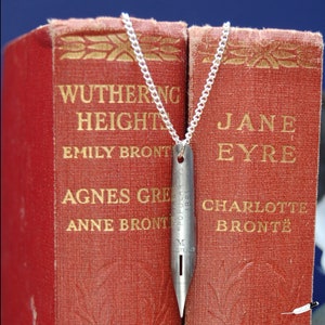 May include: A silver necklace with a vintage-style pen nib pendant. The necklace is displayed on top of two classic red hardback books, "Wuthering Heights" by Emily Brontë and "Jane Eyre" by Charlotte Brontë.
