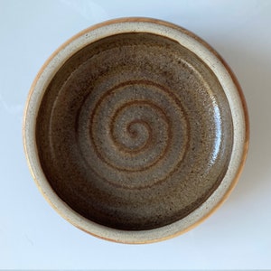 May include: A handmade ceramic bowl with a spiral pattern in the centre. The bowl has a brown interior and a light tan rim. The bowl is round and appears glazed.