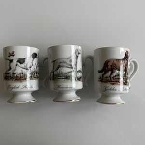 May include: Three white ceramic mugs with gold trim and illustrations of different dog breeds. The mugs are labelled "English Pointer", "Weimaraner", and "Golden Retriever".